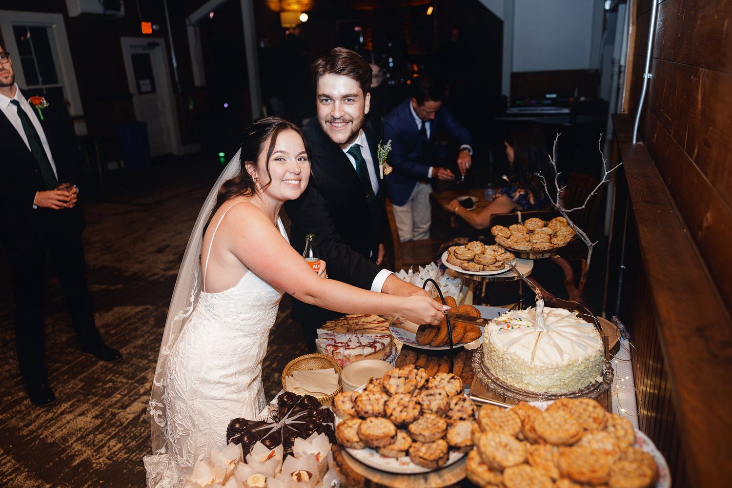 Bride and groom cutting wedding cake at dessert table filled with cookies and pastries at indoor reception.