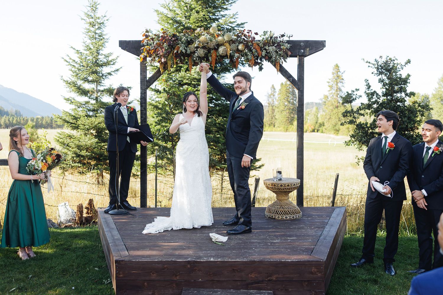 Outdoor wedding ceremony on wooden platform with couple raising hands, floral arch, wedding party, and mountain backdrop.