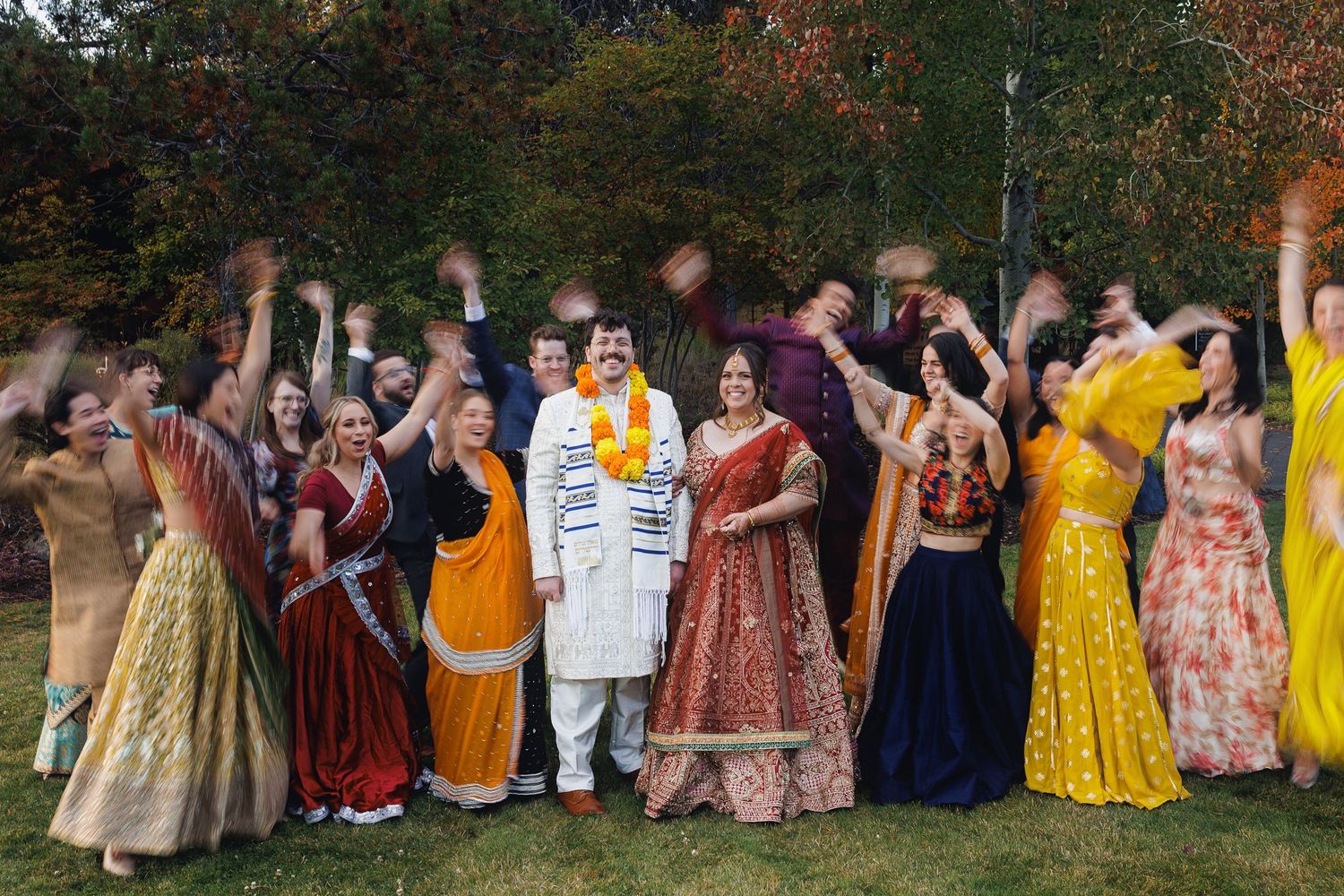 Indian wedding party celebrating outdoors in colorful traditional attire, with bride in red lehenga and groom in white sherwani.