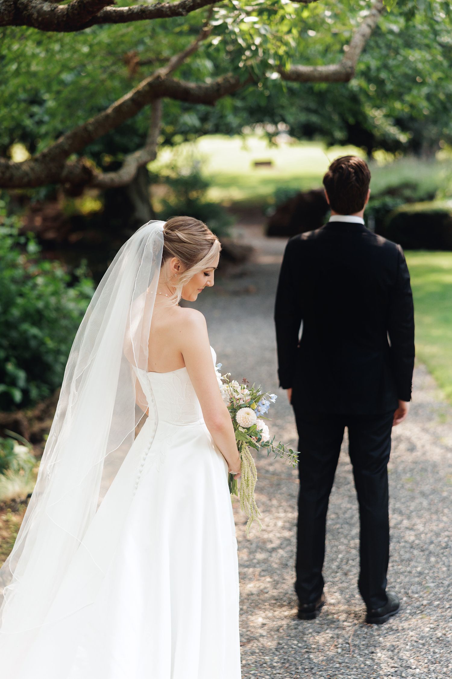 Bride in white gown and veil holding bouquet stands beside groom in black suit on a tree-lined garden path.