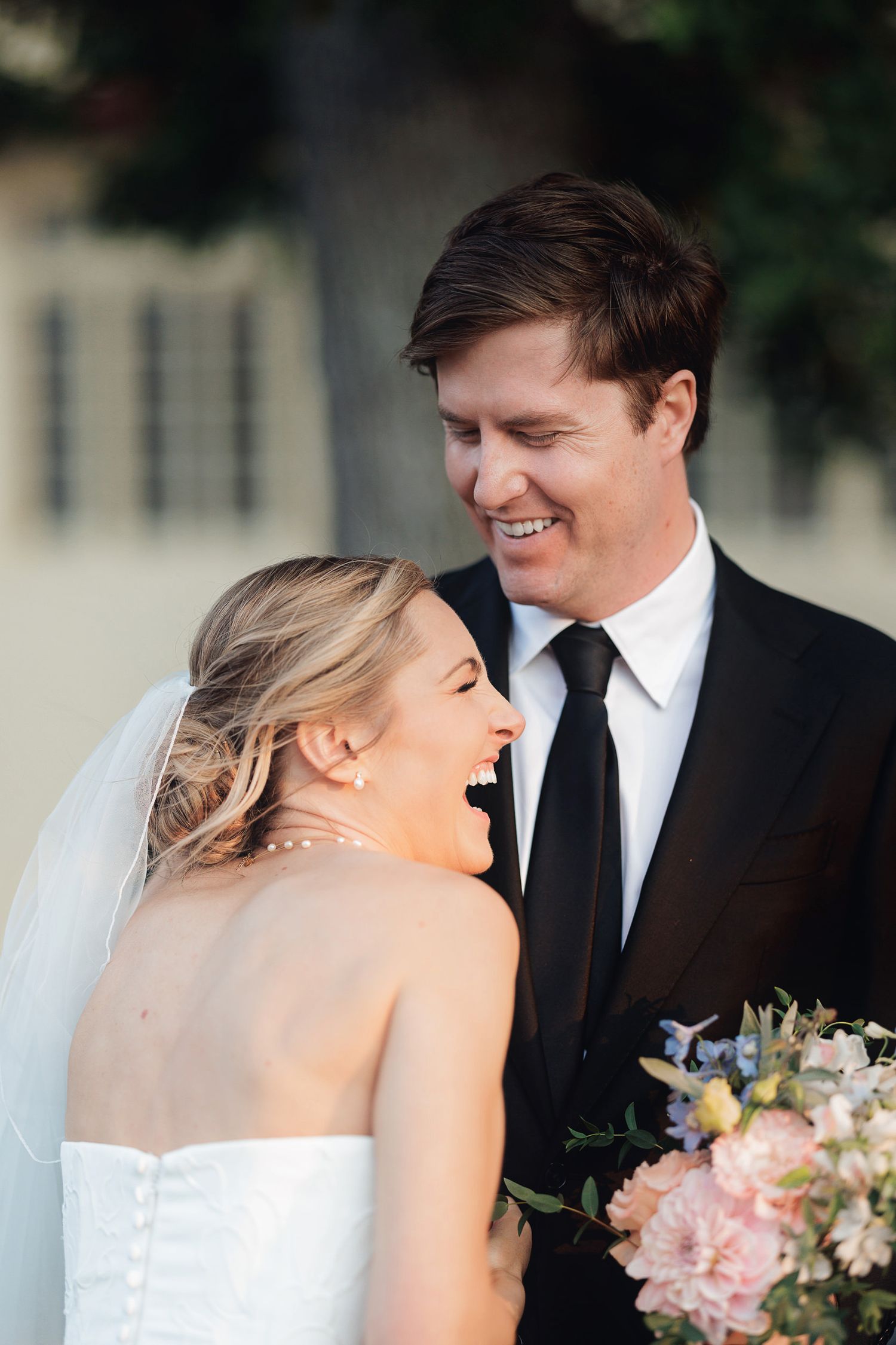 Bride and groom laughing together on wedding day, bride in strapless gown holding pink floral bouquet, groom in black suit.