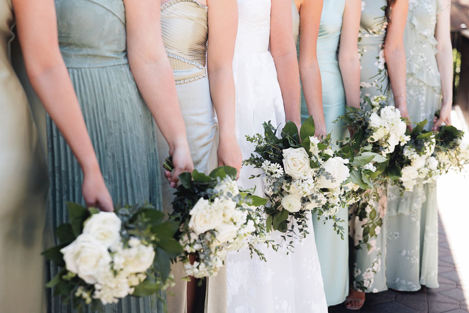 Row of bridesmaids in mismatched sage dresses holding white rose and greenery bouquets beside bride.