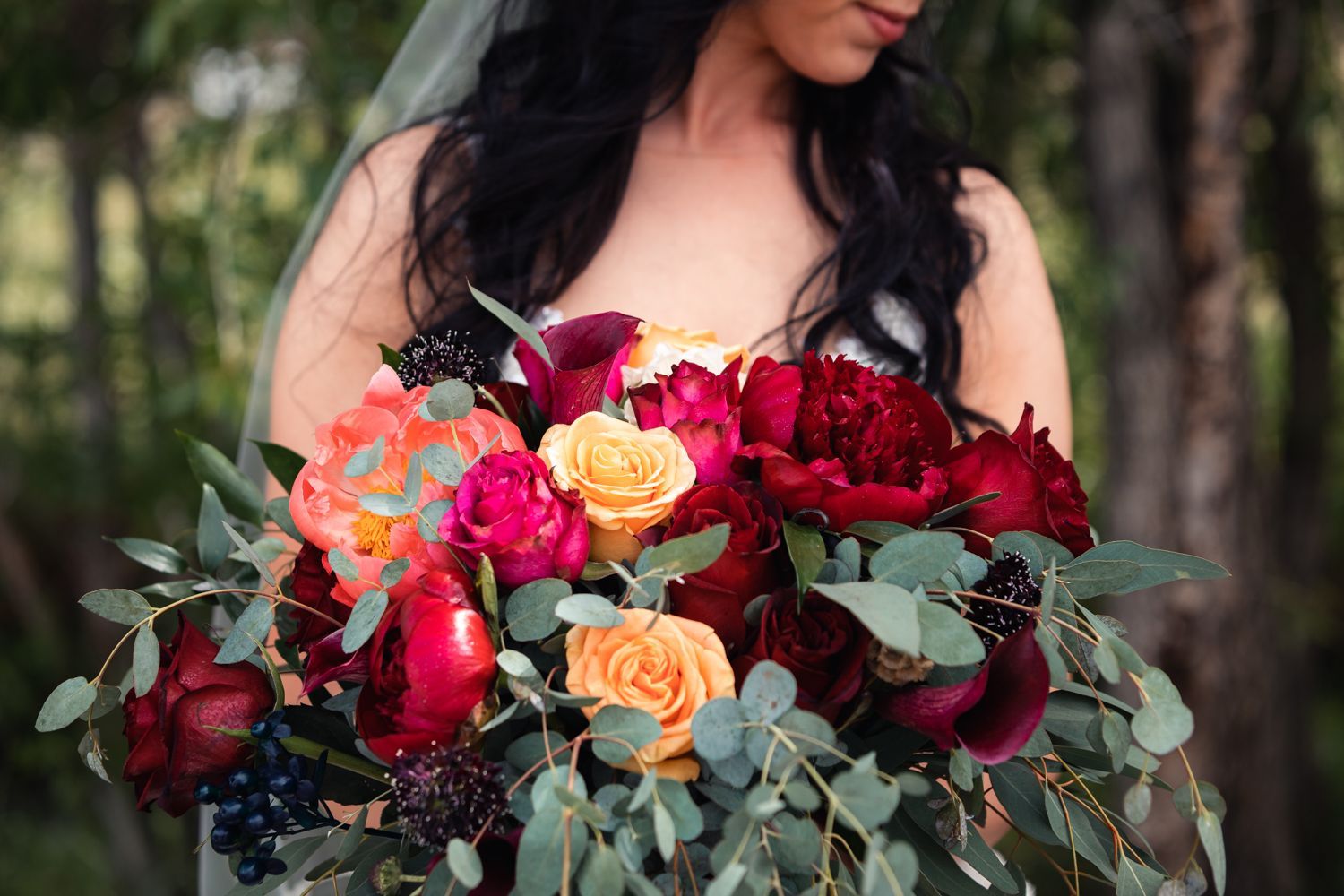 A stunning bridal bouquet featuring deep red and peach roses with eucalyptus greenery against a forest backdrop.