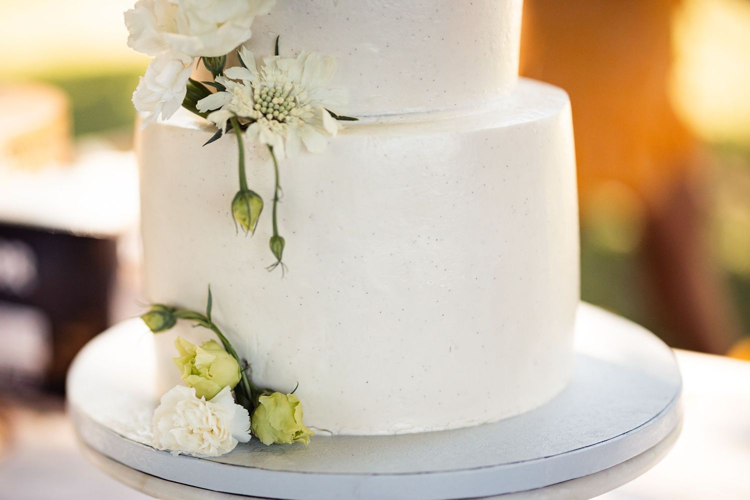 Elegant white wedding cake decorated with delicate white and pale green flowers on a silver cake stand.
