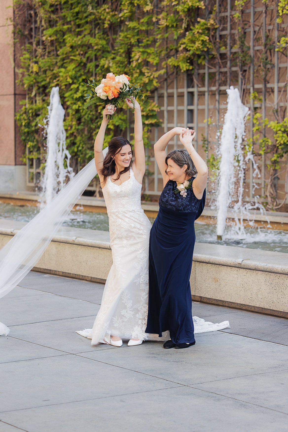 Two people in formal dresses celebrate joyfully in front of a fountain with ivy-covered walls.
