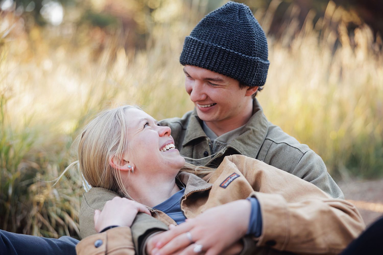 Smiling couple in rust and olive jackets embrace outdoors, the woman laughing in his lap as he wears a gray beanie amid golden autumn grass.