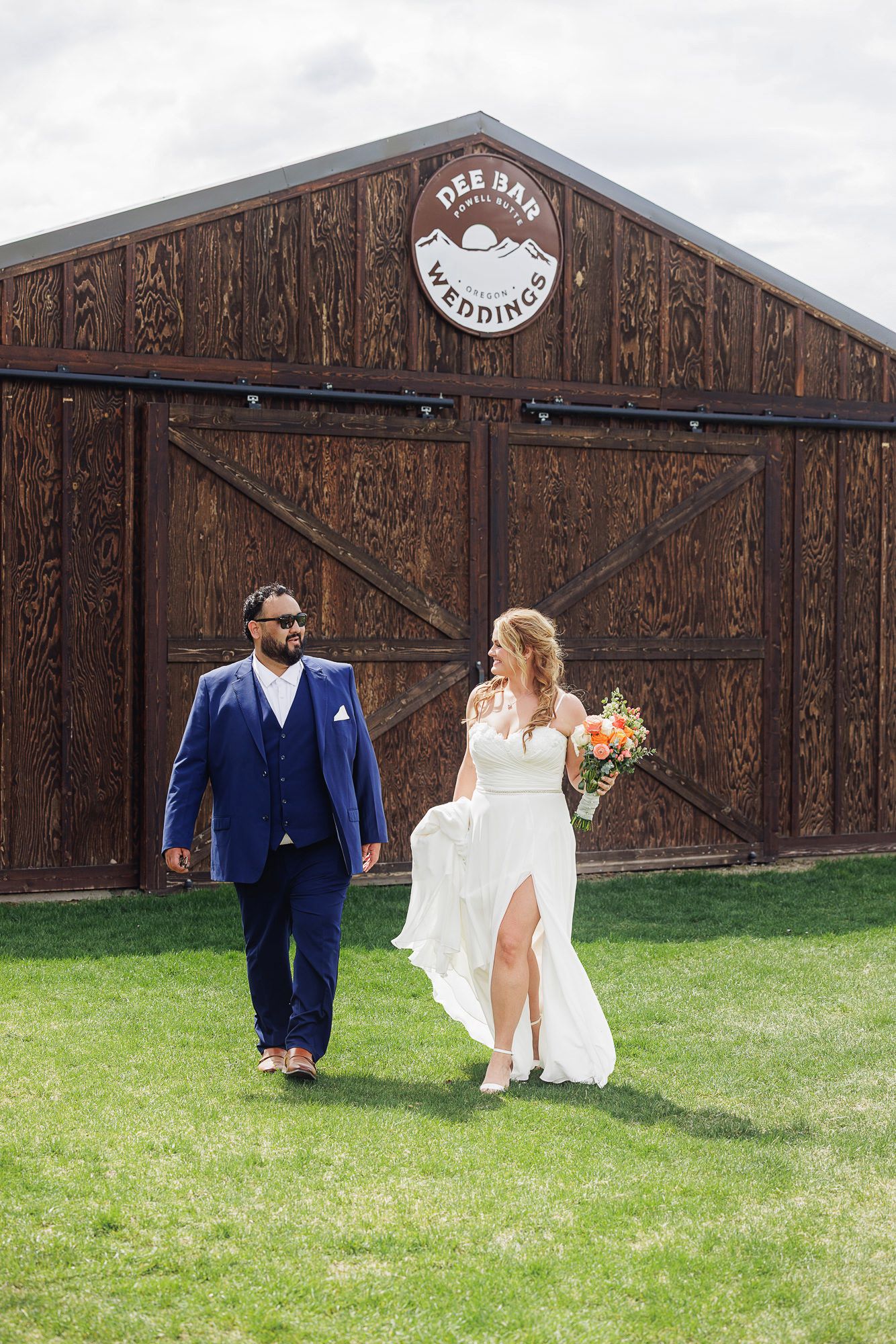 Bride and groom walking on green grass in front of Dee Bar Weddings rustic brown barn venue on cloudy day.