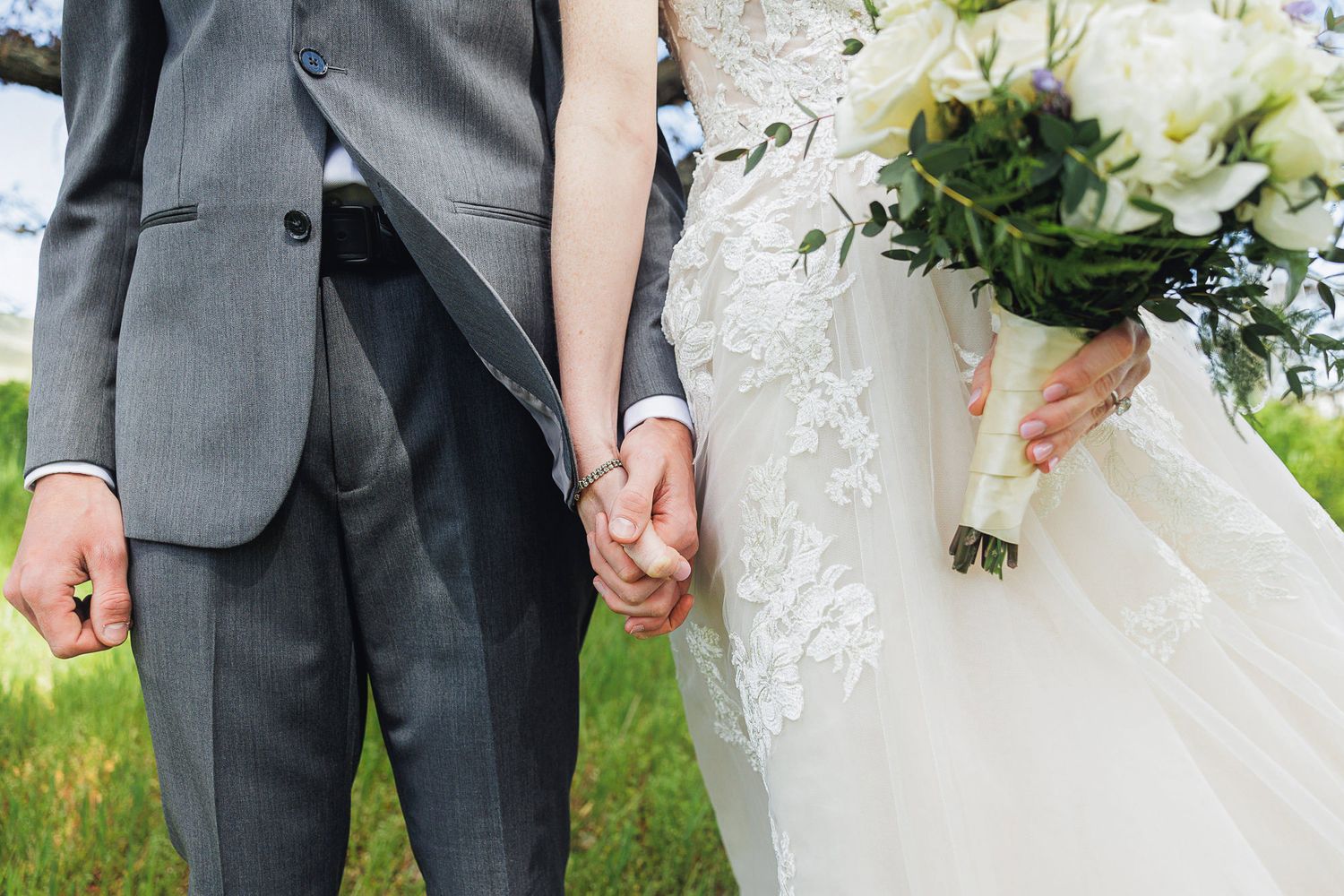 Newlywed couple holding hands, bride in lace wedding gown with bouquet, groom in gray suit, outdoor ceremony.