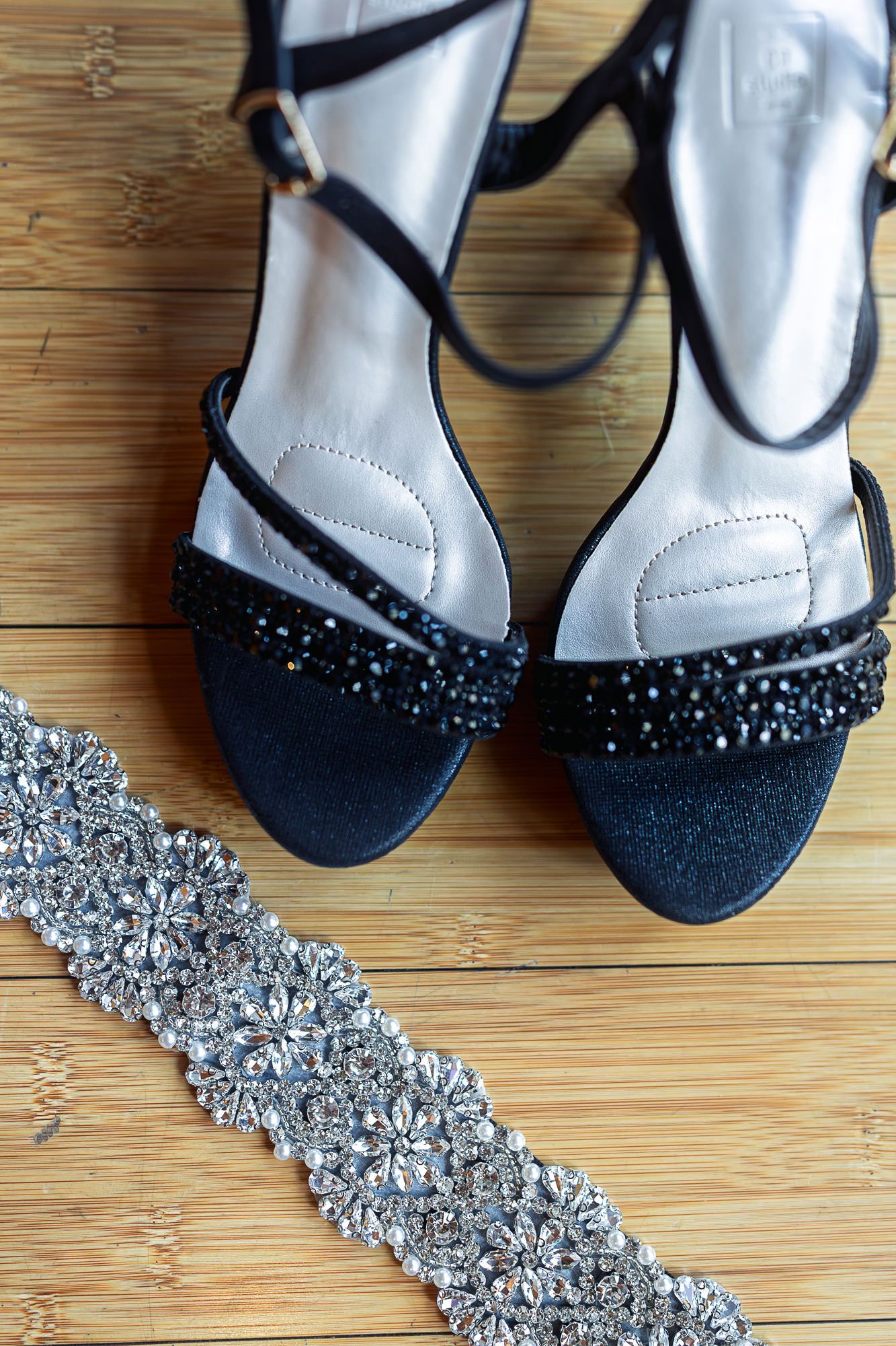 Black rhinestone embellished heeled sandals displayed on wooden floor beside a crystal beaded bridal sash belt.