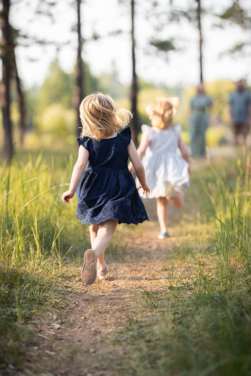 Two barefoot children in summer dresses run along a dirt path through tall grass and trees on a sunny afternoon.