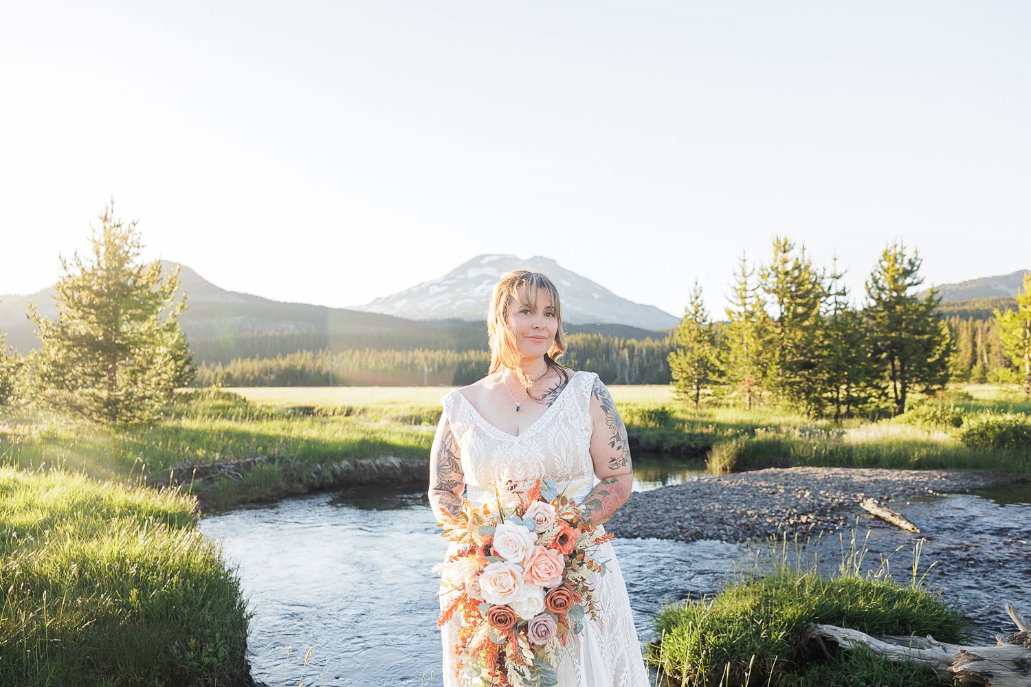 Person in a white dress holding a bouquet stands by a flowing stream with mountains and evergreen trees in the background.