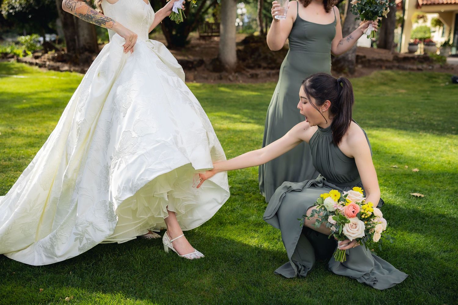 A bridesmaid in a sage green dress helps adjust a flowing white wedding gown outdoors on a sunny lawn.
