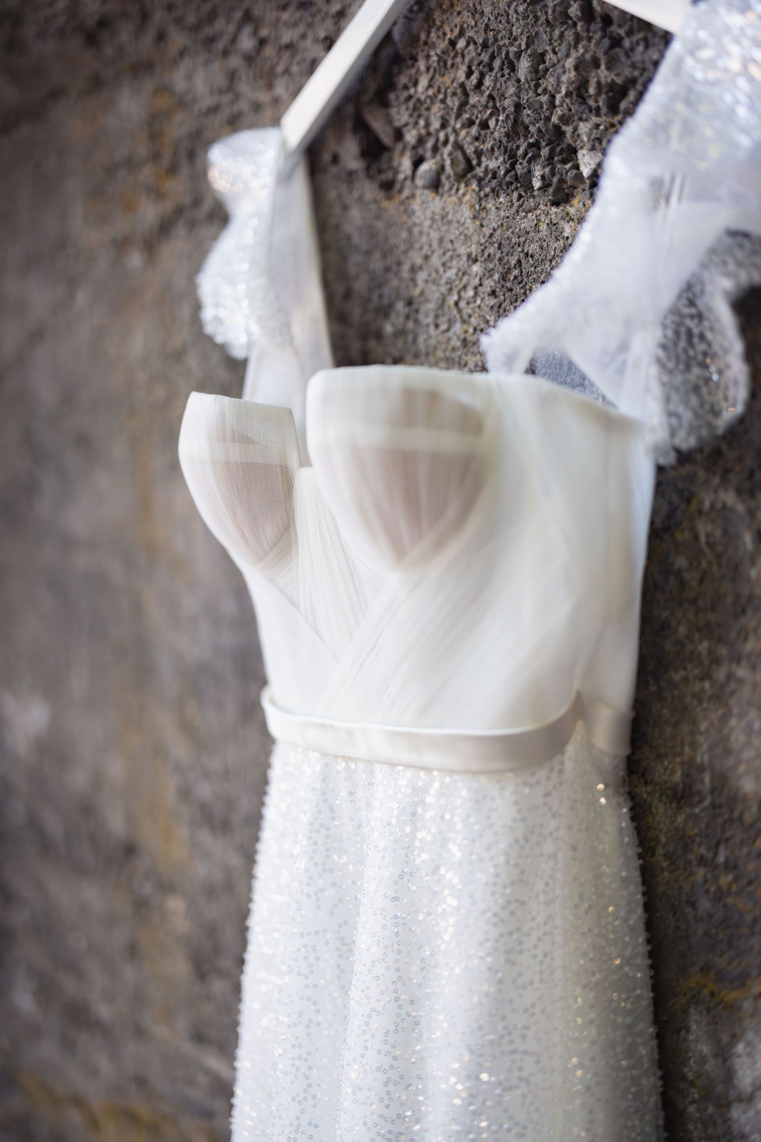 A white strapless wedding dress with a glittering sparkly skirt hangs against a textured stone wall.