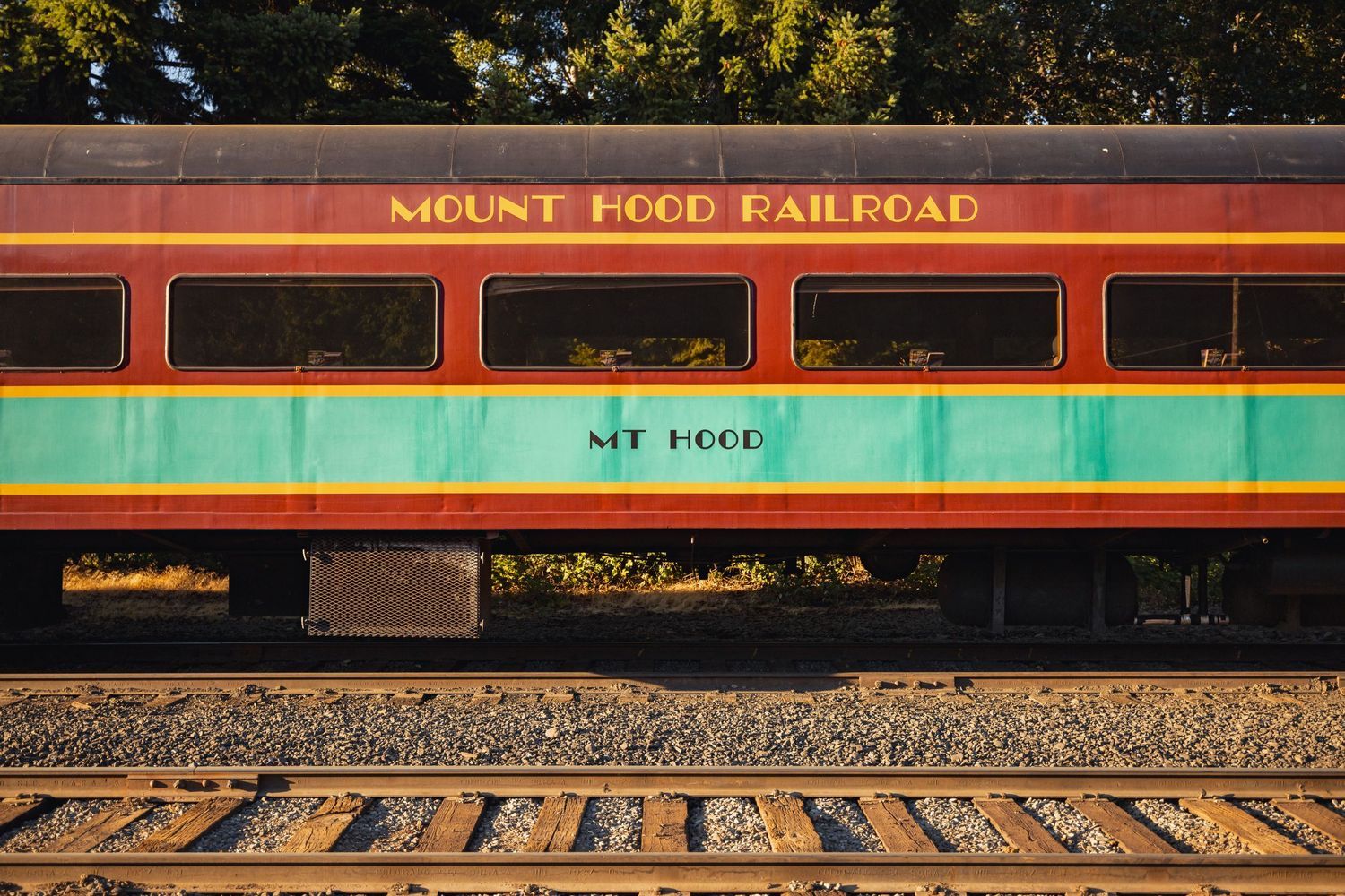 A vintage red and turquoise Mount Hood Railroad passenger car sits on train tracks during golden hour.