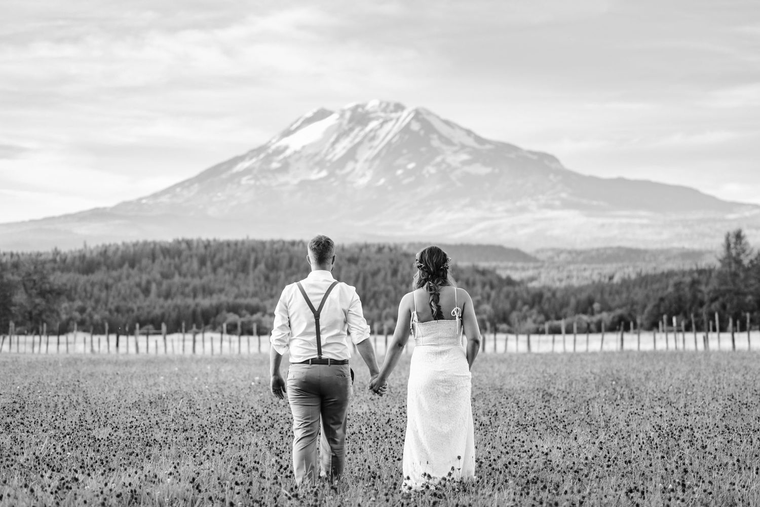 Black and white image of a couple holding hands while facing a snow-capped mountain in a vast field.