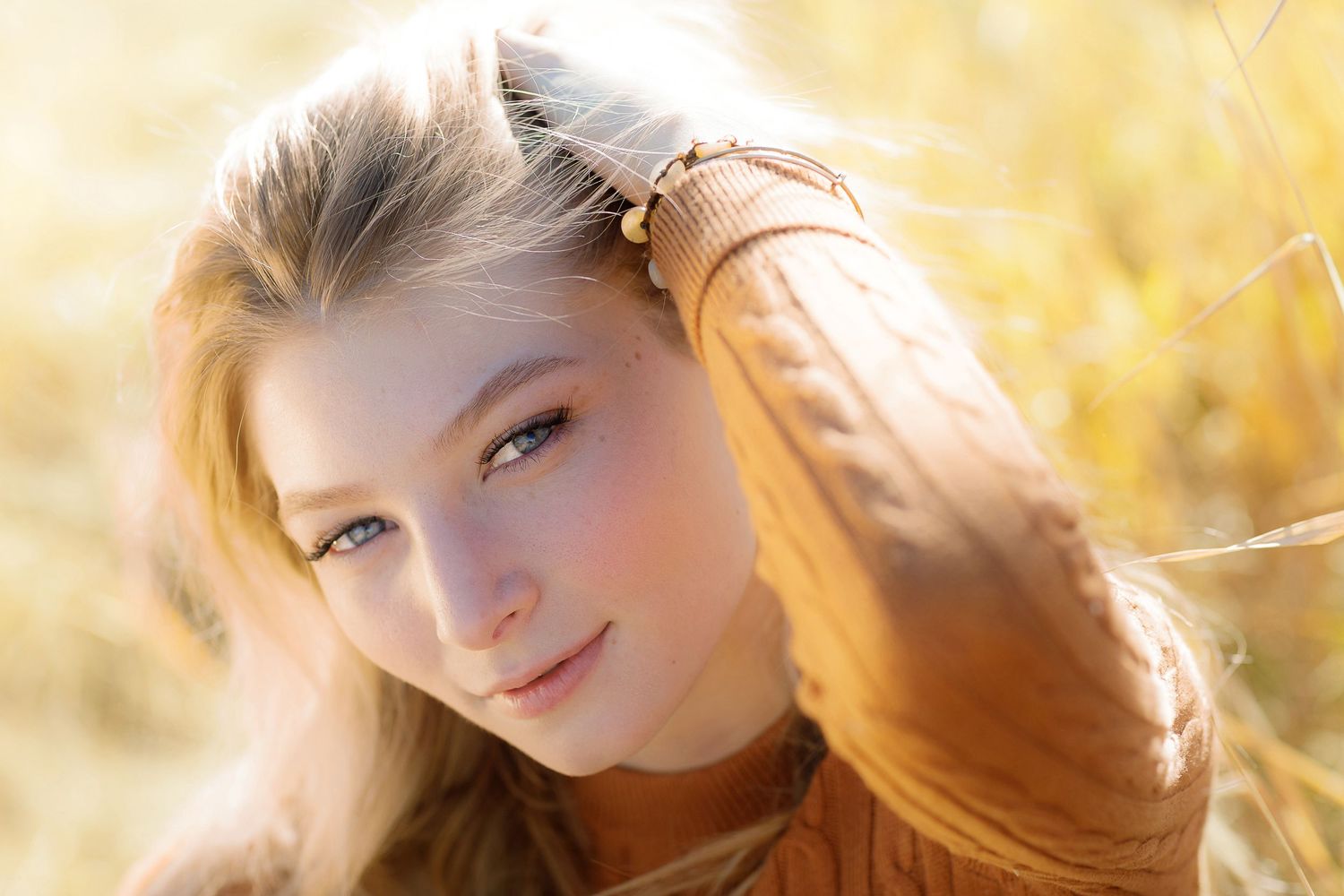 A young portrait subject with long blonde hair in a brown sweater smiling in golden sunlight outdoors.