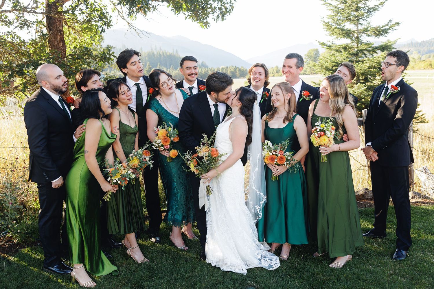 Wedding party in green dresses and black suits kiss bride outdoors with mountain backdrop and fall bouquets.