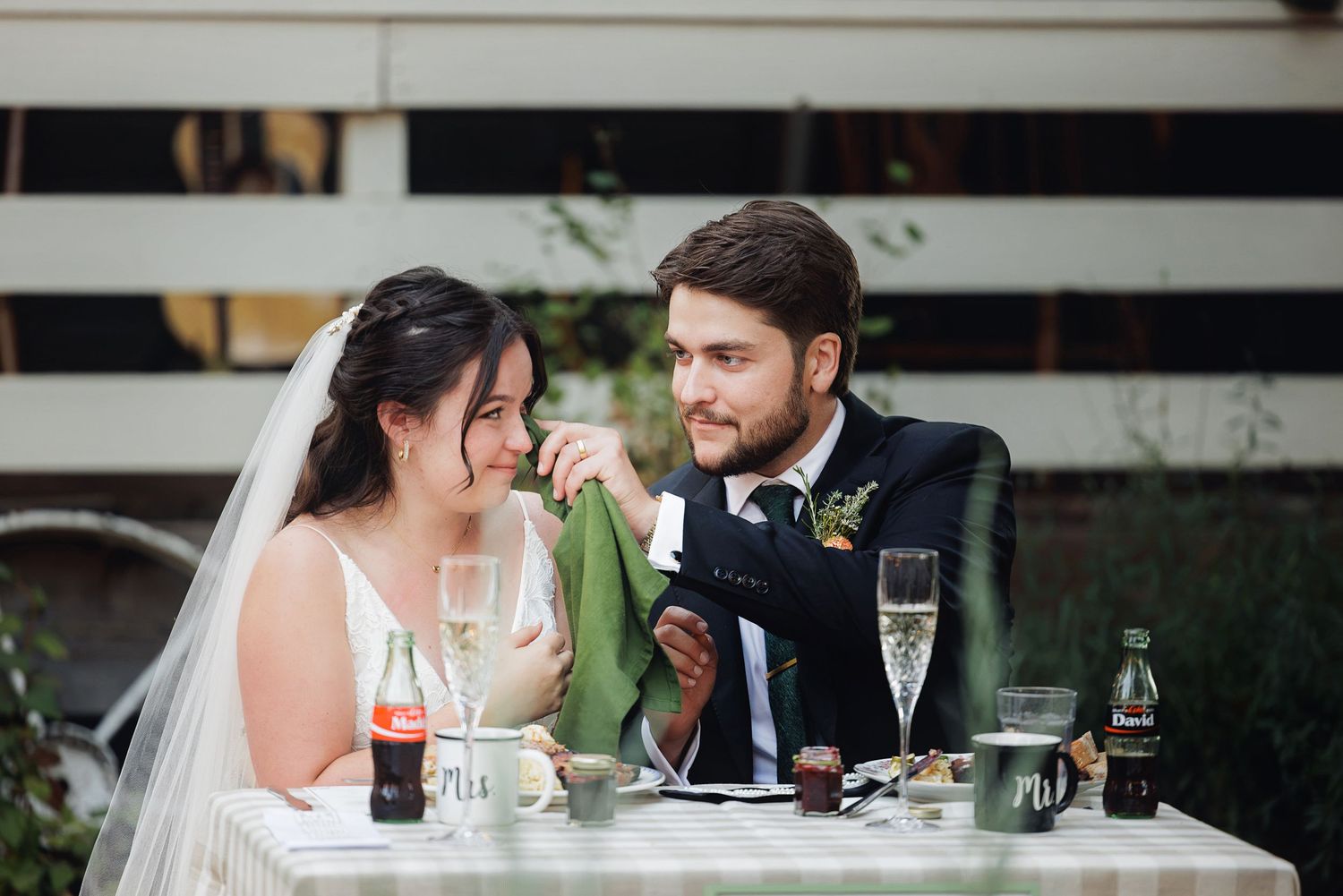 Bride and groom share a tender moment at outdoor wedding reception table with champagne glasses and Coca-Cola bottles.