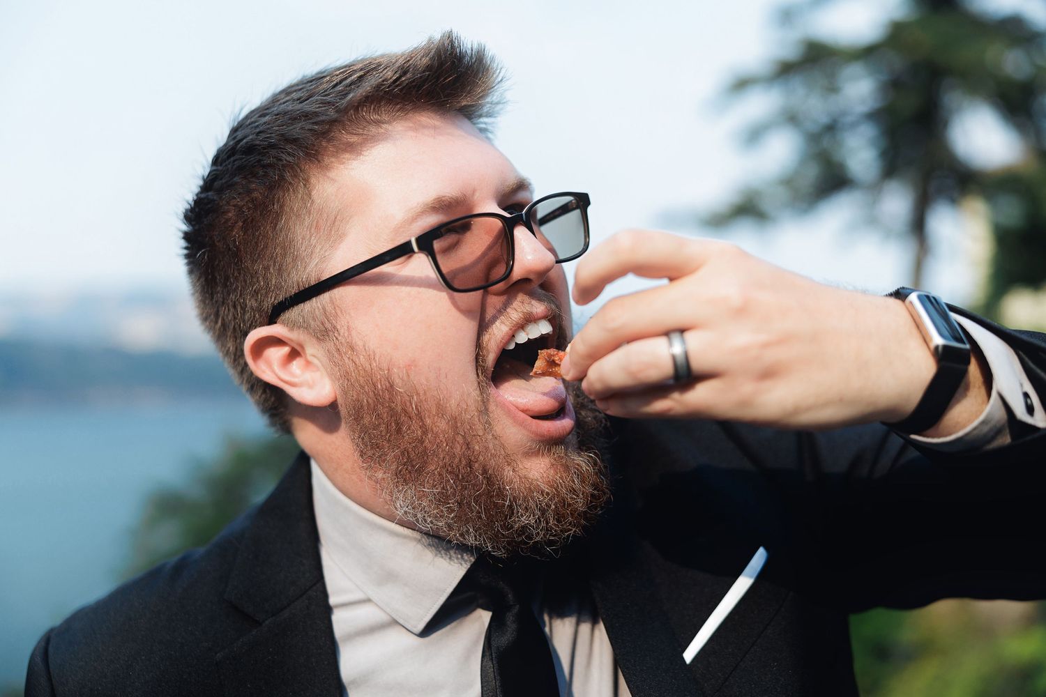 Bearded man in suit and glasses eating food at outdoor event with scenic background.
