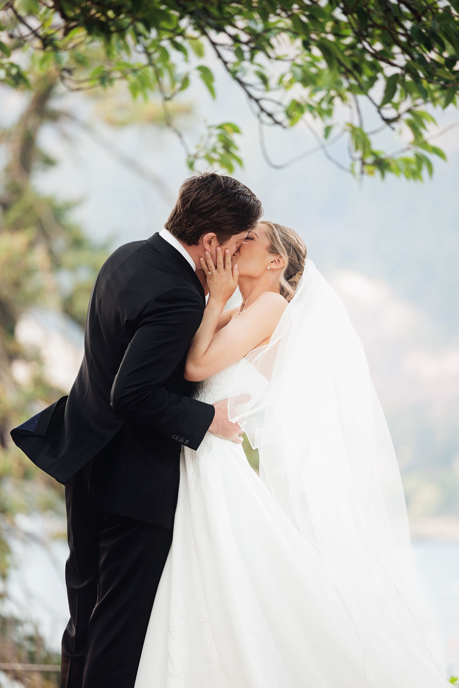 Bride and groom share first kiss in outdoor wedding ceremony, bride in white gown with veil, groom in black suit.