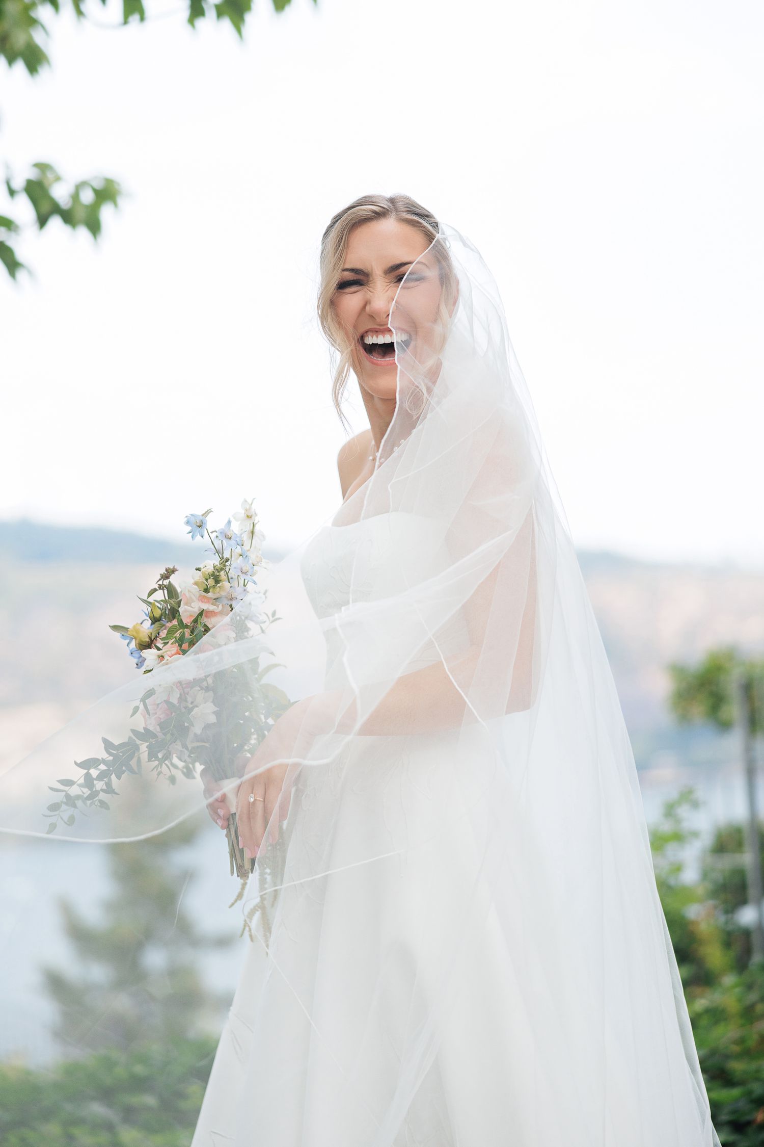 Joyful bride in white gown and veil laughing outdoors, holding a pastel bouquet with greenery on her wedding day.