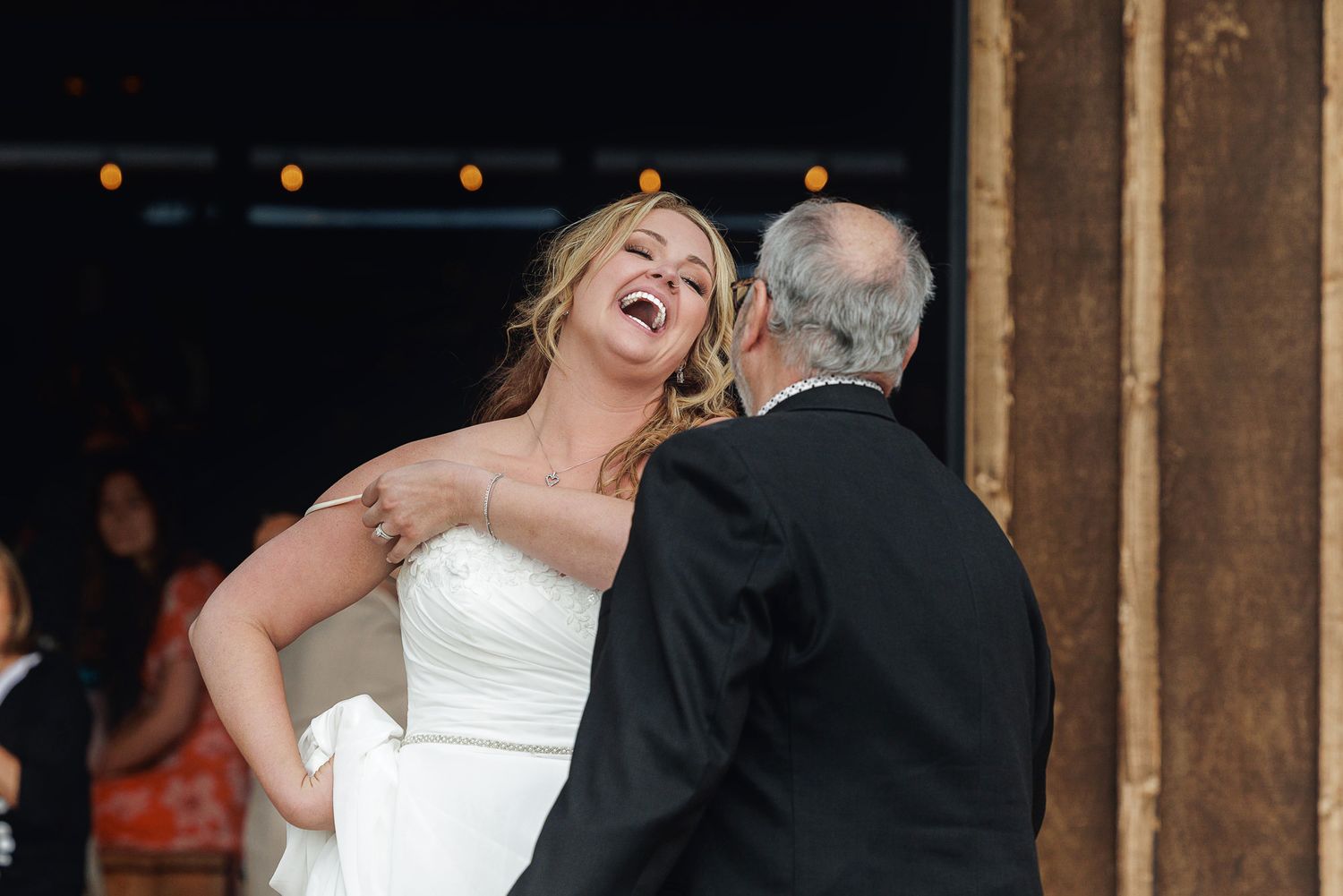Bride in white gown laughing joyfully during father-daughter dance at rustic wedding reception.