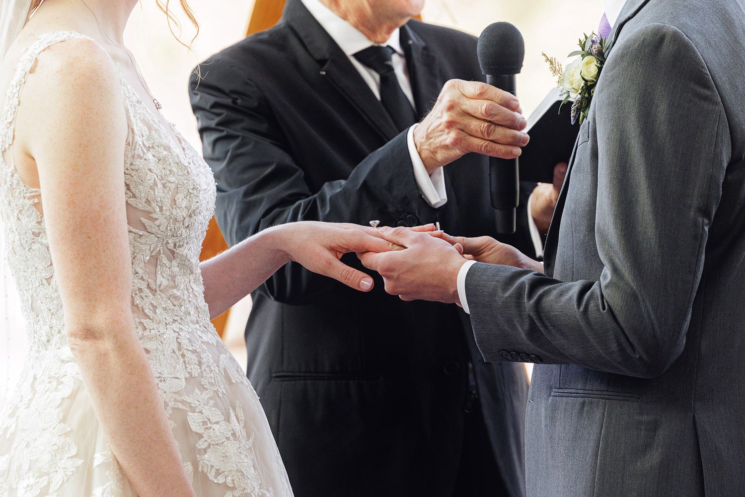 Officiant performing wedding ring exchange ceremony between bride in lace gown and groom in gray suit.