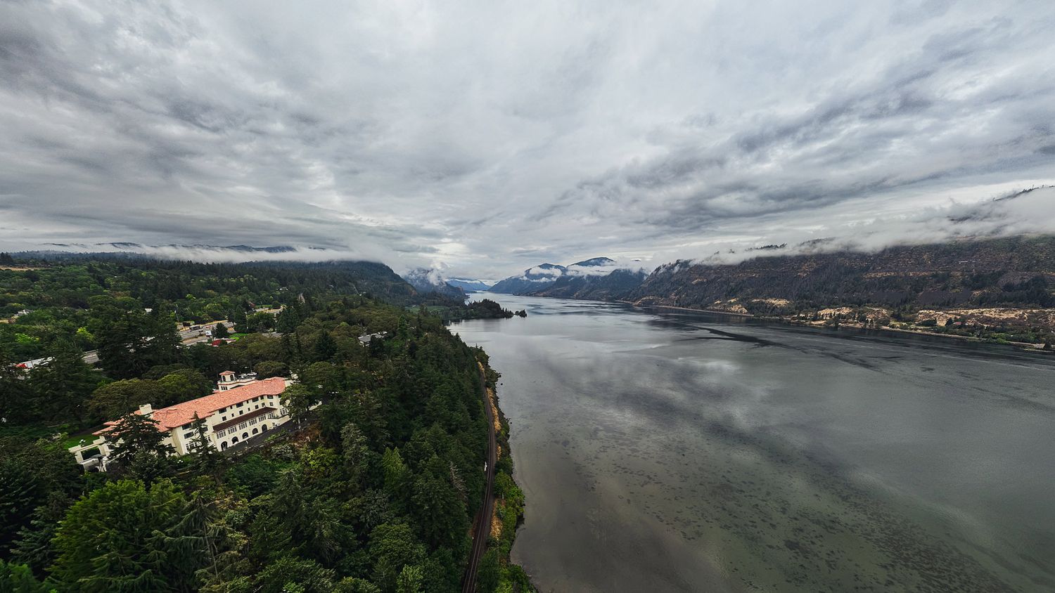 Aerial view of a historic mansion on a cliff overlooking the Columbia River Gorge under dramatic cloudy skies.