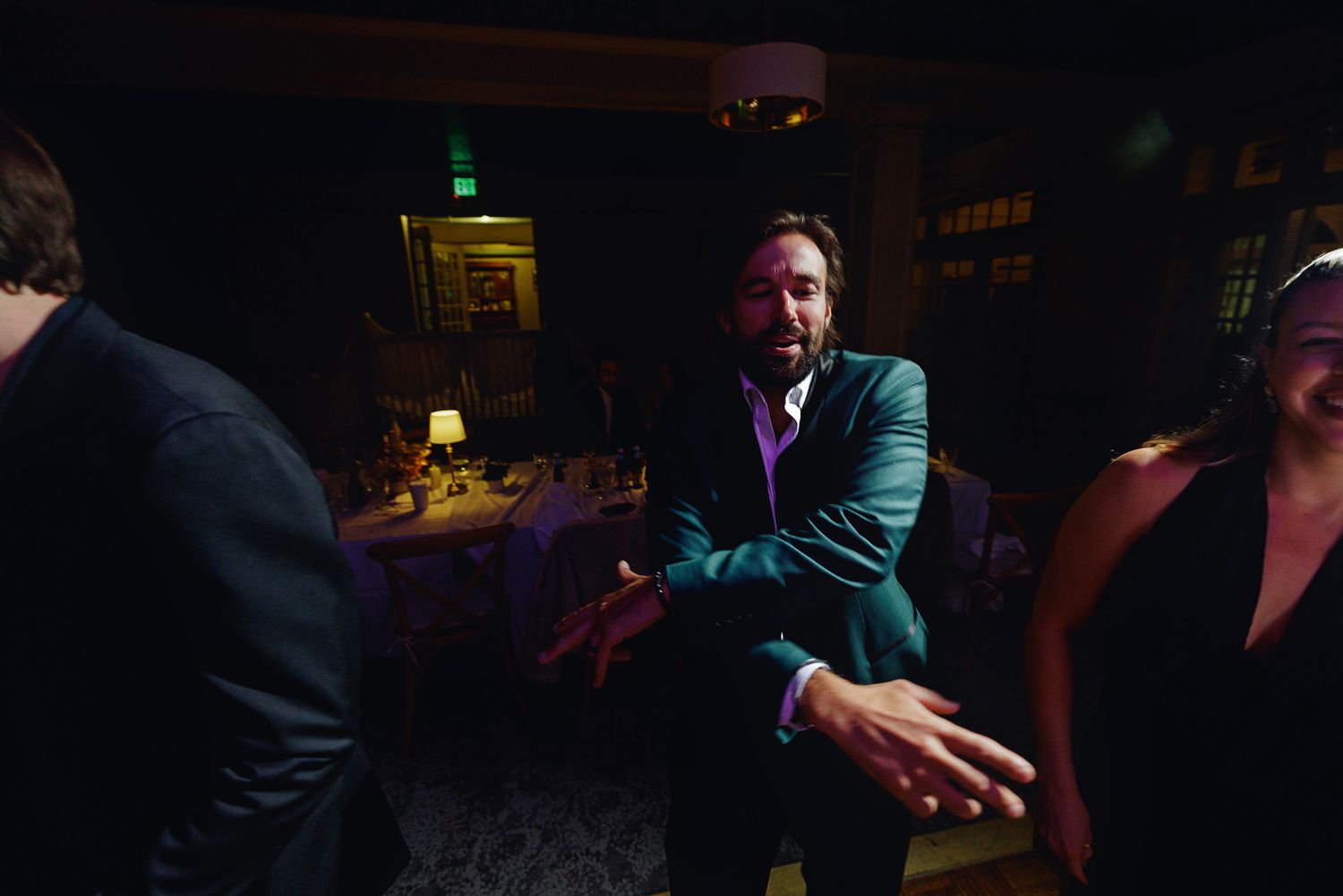 Man in teal suit jacket dancing at dimly lit elegant event venue with other guests.