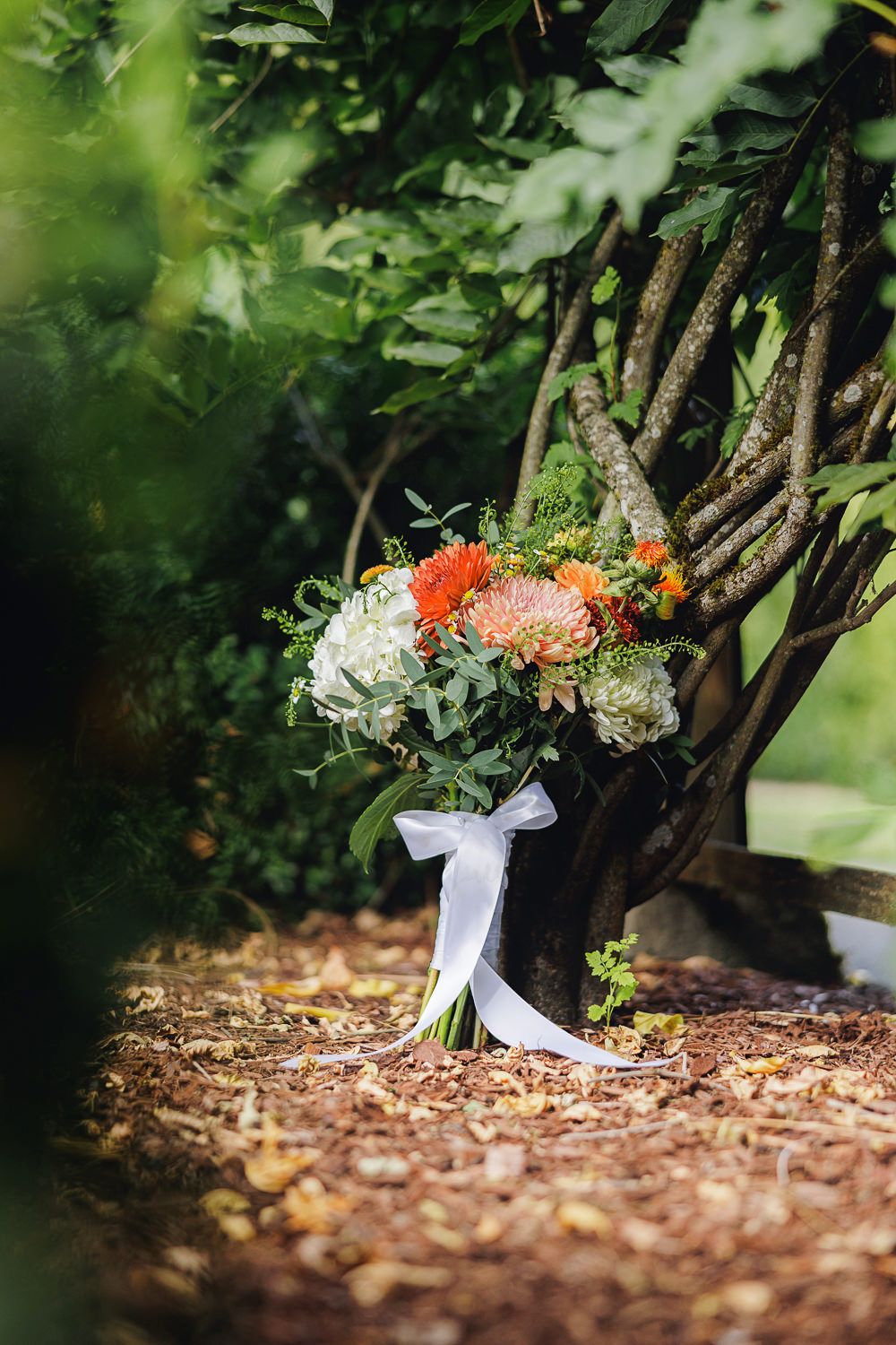 A rustic wedding bouquet with white and coral flowers tied with a white ribbon rests against tree branches in a garden.