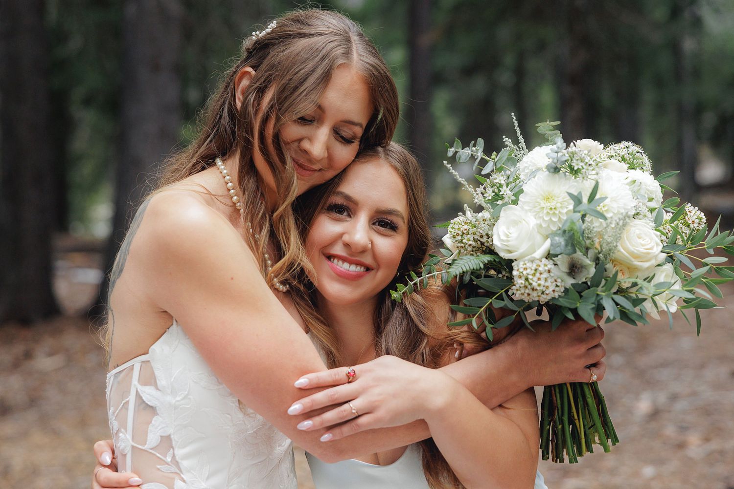 Two people in wedding attire share a joyful embrace while holding a beautiful white floral bouquet in a forest setting.