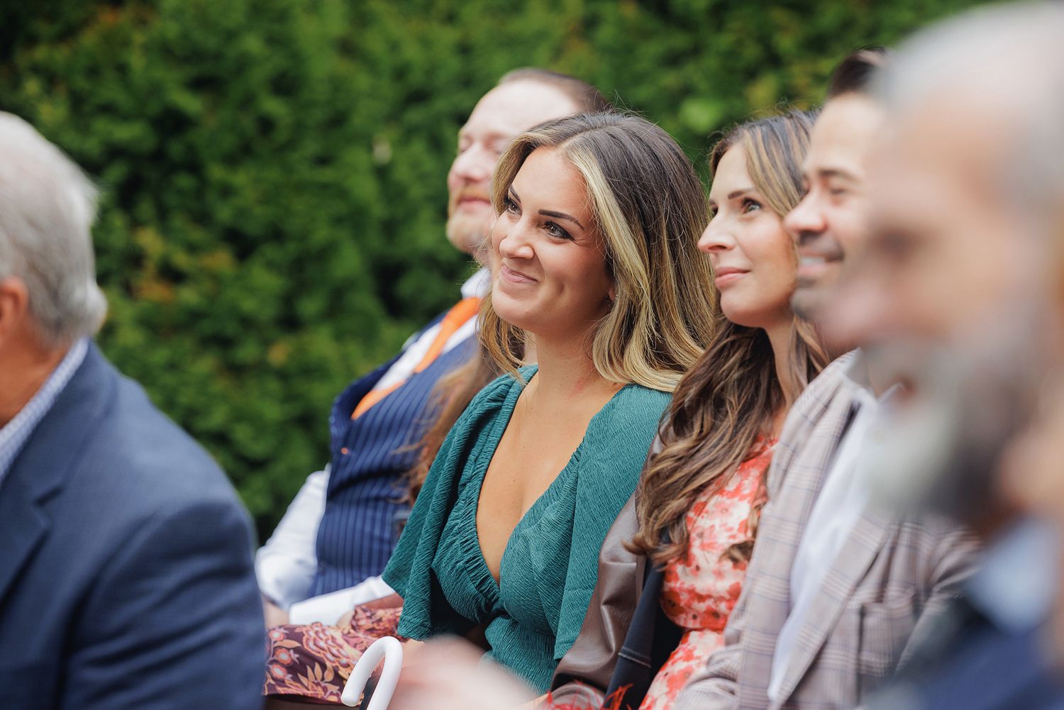 Wedding guests seated outdoors watching ceremony with greenery in background.
