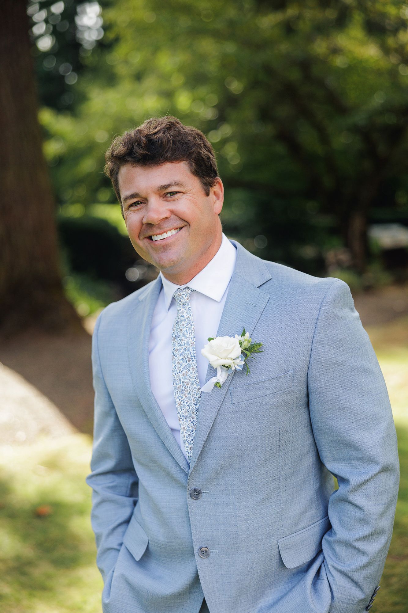 A person in a light gray suit and patterned tie poses outdoors against a blurred green natural background.