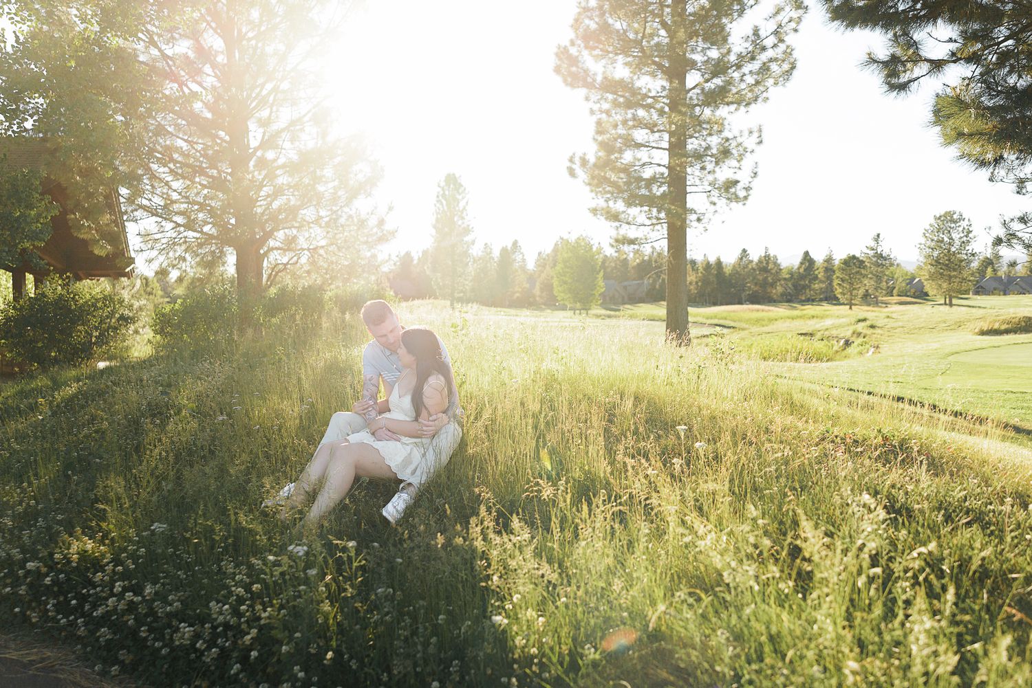 Couple sitting together in sunlit meadow surrounded by tall green grass and trees during golden hour outdoor photo session.