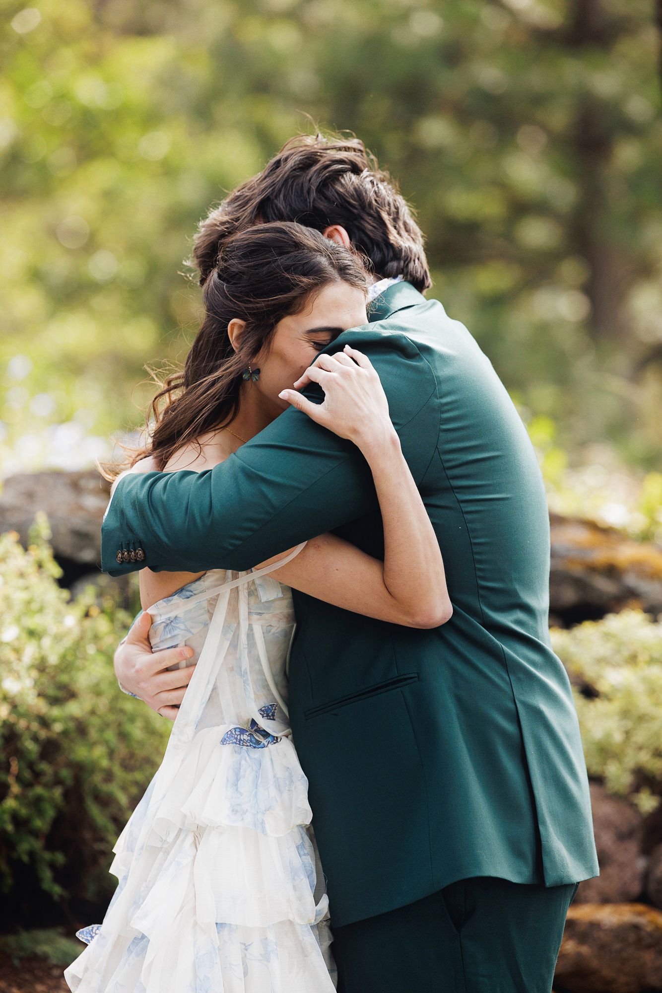 A bride and groom share an emotional embrace outdoors, with the groom wearing a green suit and bride in a ruffled white gown.