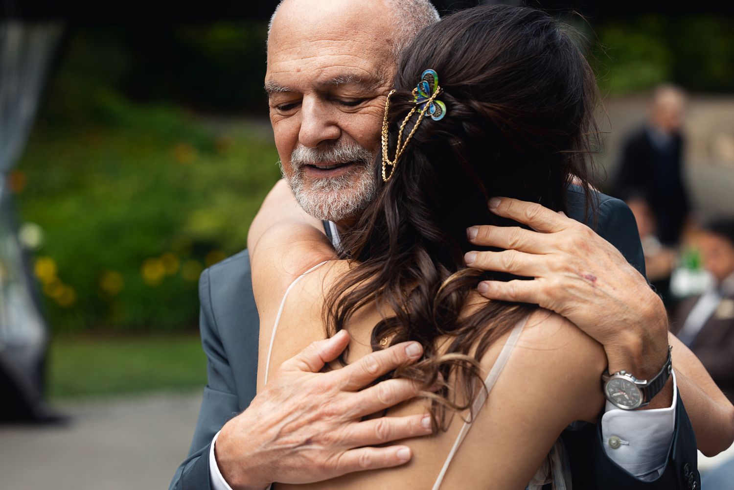 Elderly man with gray beard sharing an emotional hug with a bride in a strapless gown at an outdoor wedding ceremony.