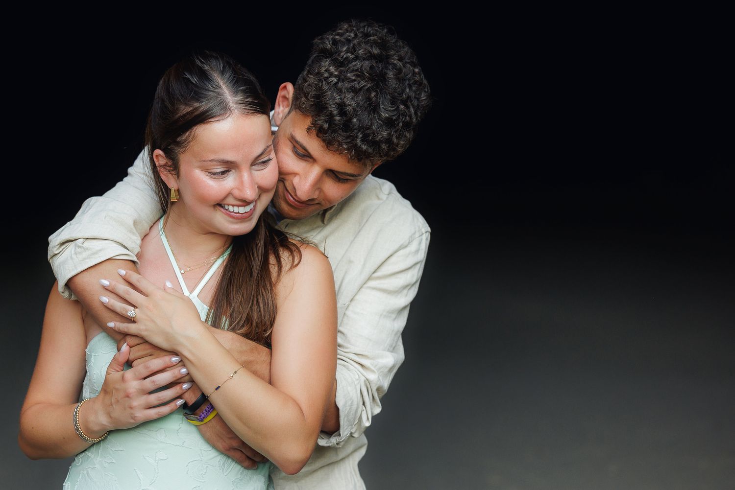 Young couple embracing warmly, smiling woman in mint dress with man in beige suit on dark background.