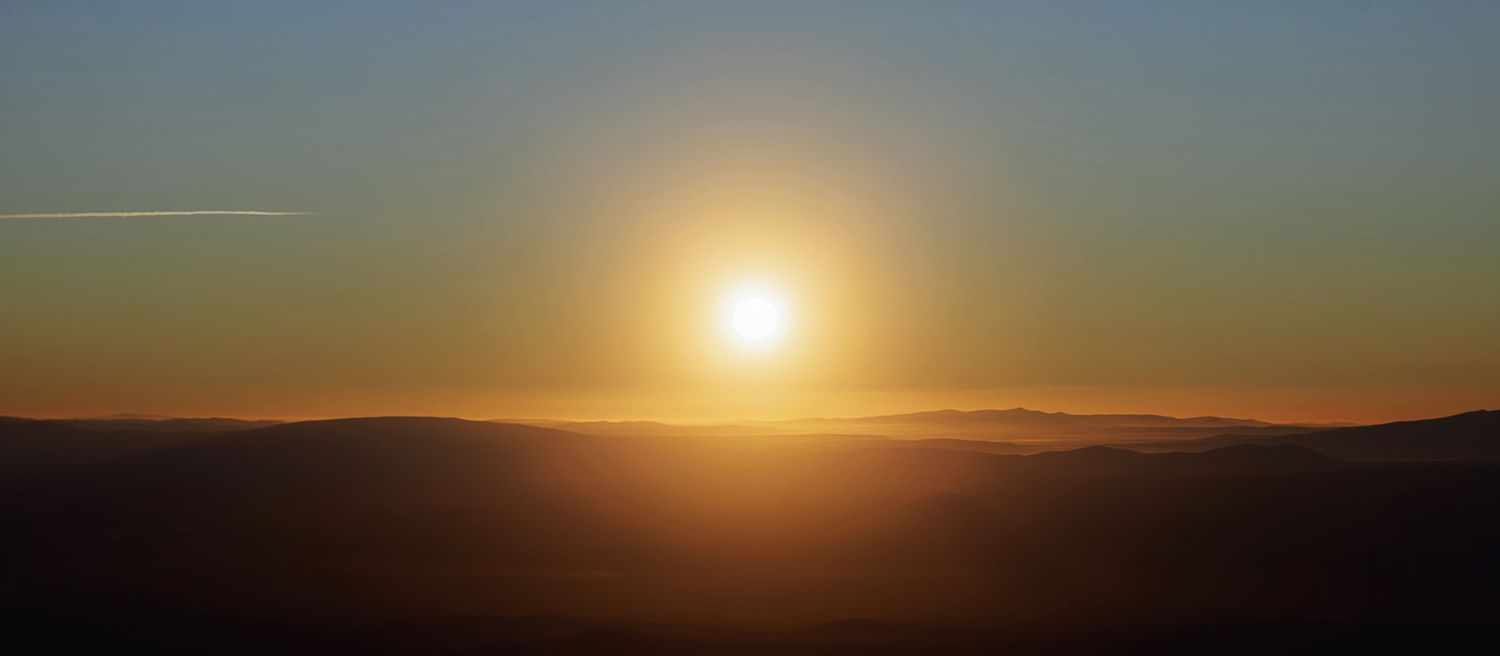 Golden sun glowing over layered mountain silhouettes at sunrise or sunset, casting warm orange light across a hazy sky.