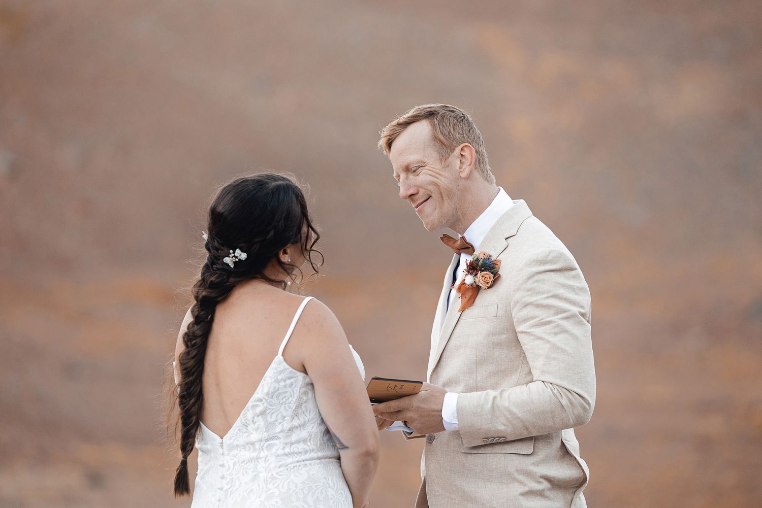 Smiling groom in a beige suit with a floral boutonniere reads vows to his bride, who wears a lace wedding dress and a long braid, at an outd