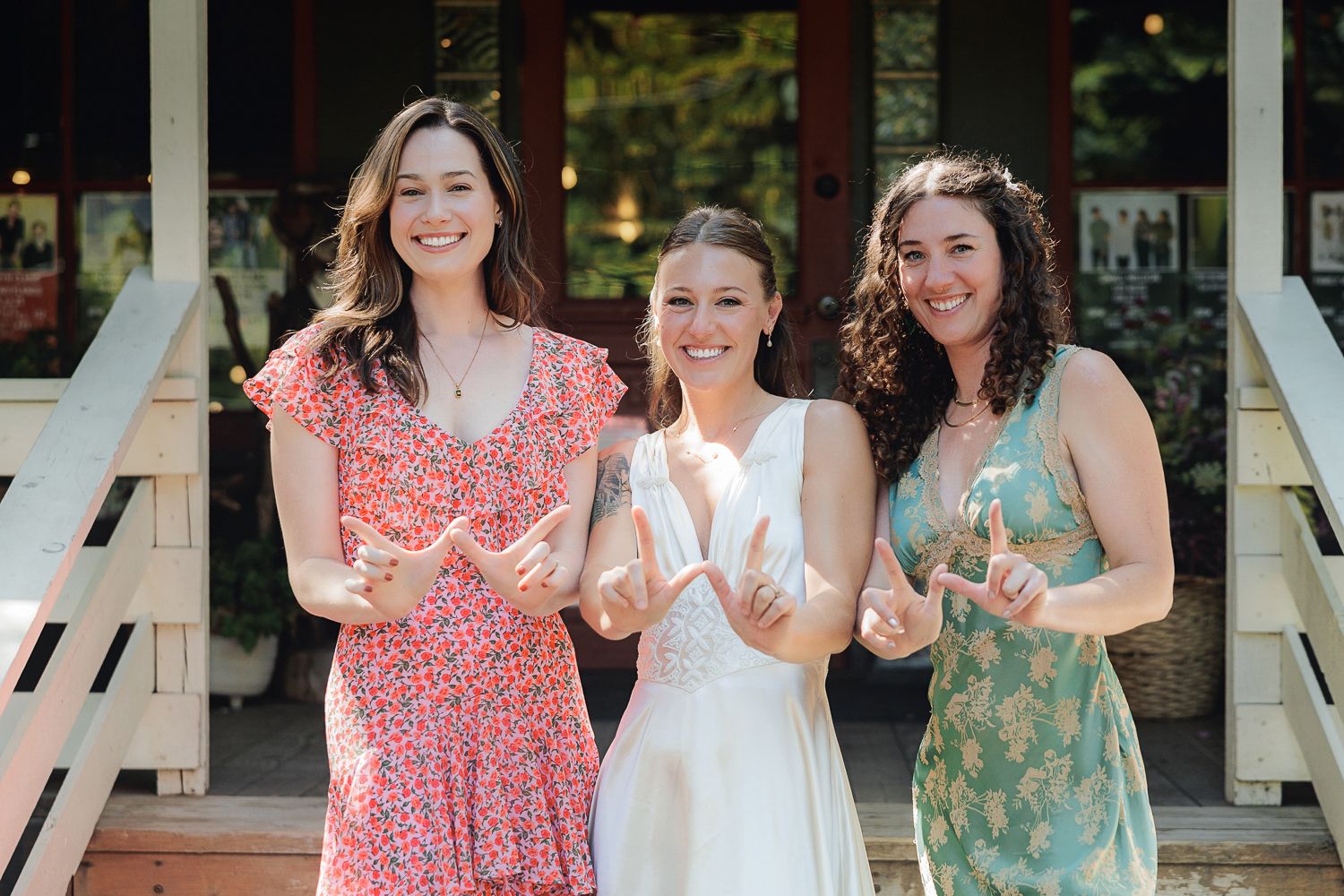 Three women in summer dresses stand together smiling on wooden porch steps of a building.
