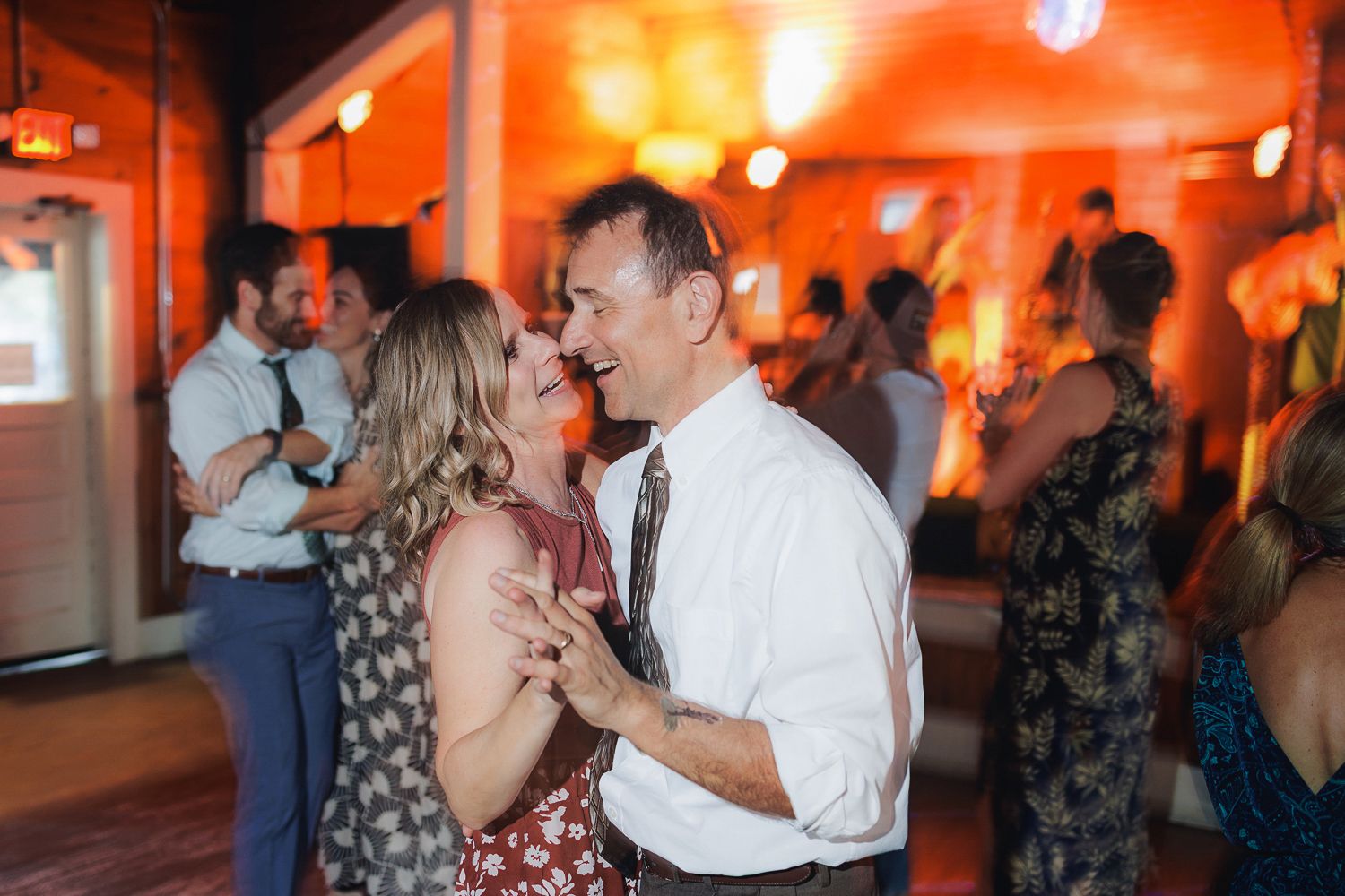 Dancing couples share intimate moments in an orange-lit reception venue space.