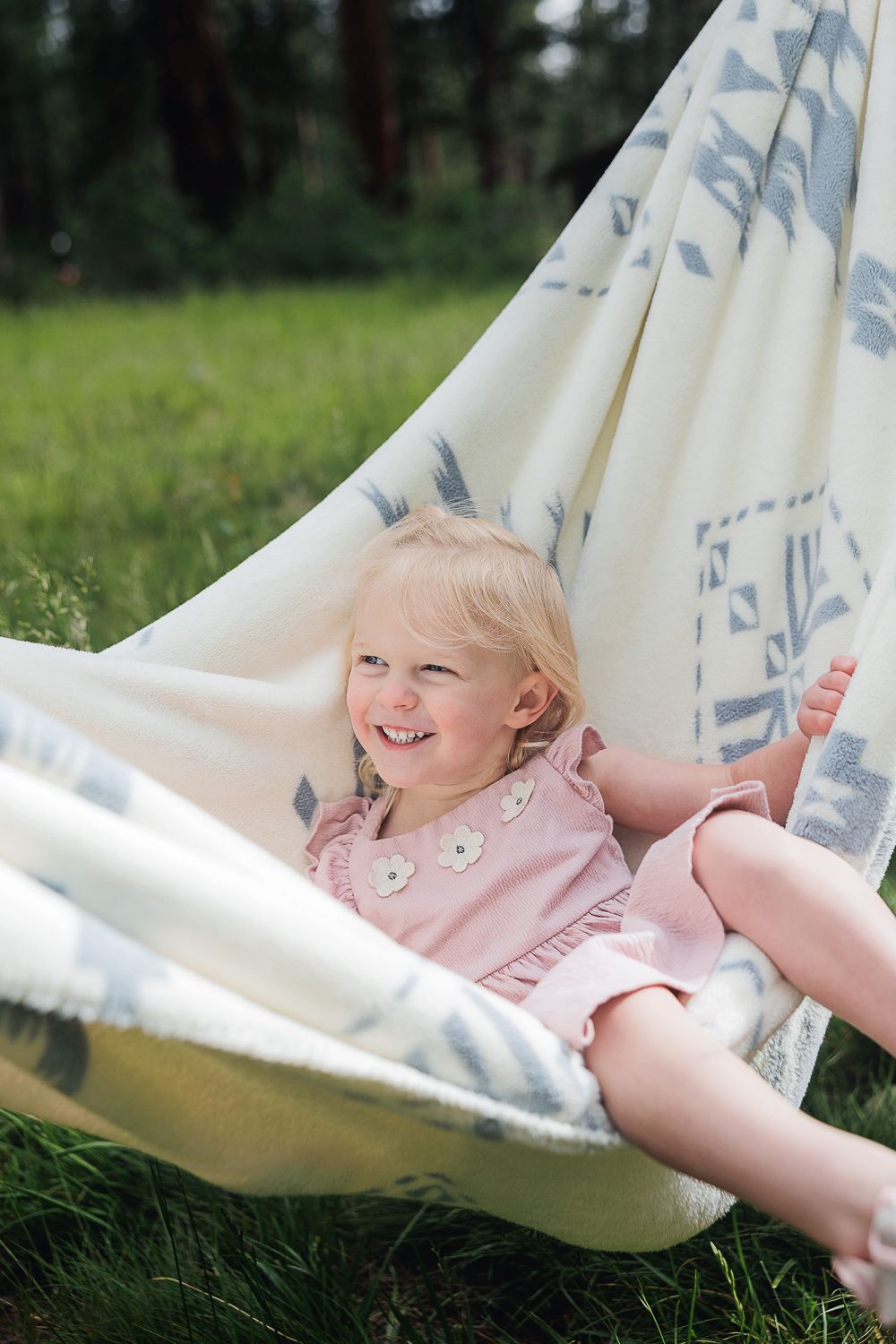 Smiling toddler wearing pink dress lounges in white patterned hammock on sunny day.