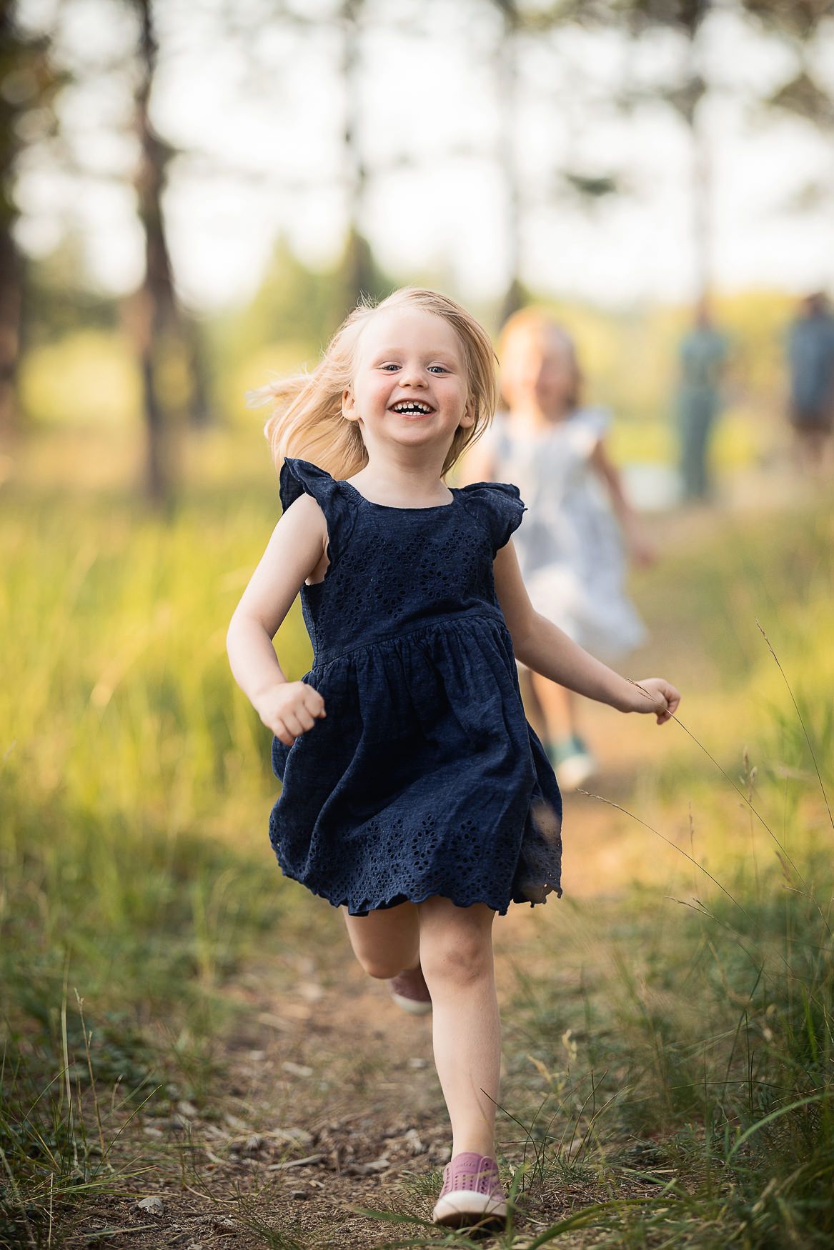 A young child in a navy blue dress runs joyfully along a dirt path through tall grass on a sunny day.