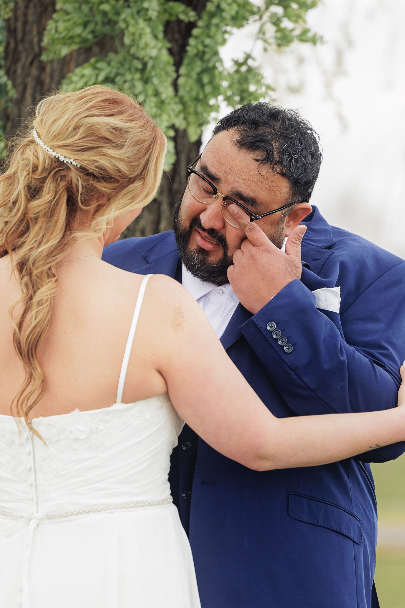 Emotional groom in blue suit wiping tears during wedding first look with blonde bride in white dress outdoors.