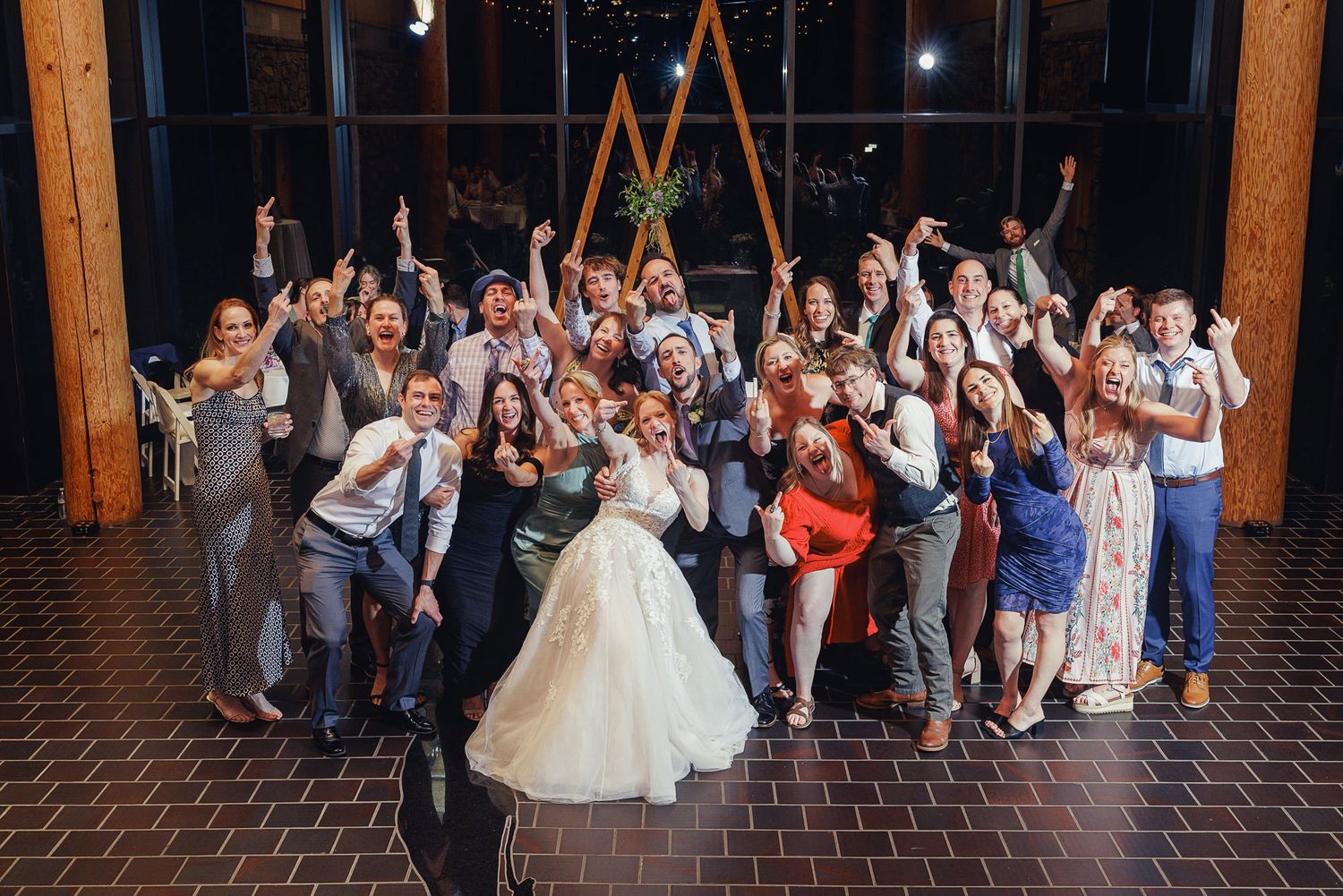 Bride and groom pose with joyful wedding guests cheering in a warmly lit reception venue.