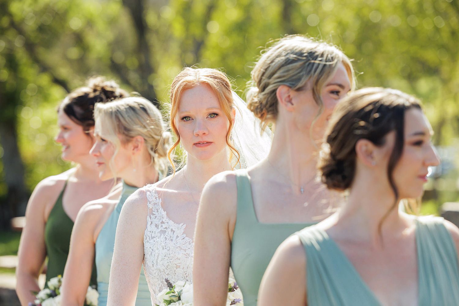 Bridesmaids in sage green dresses stand outdoors among spring trees, with the bride in white lace at center.
