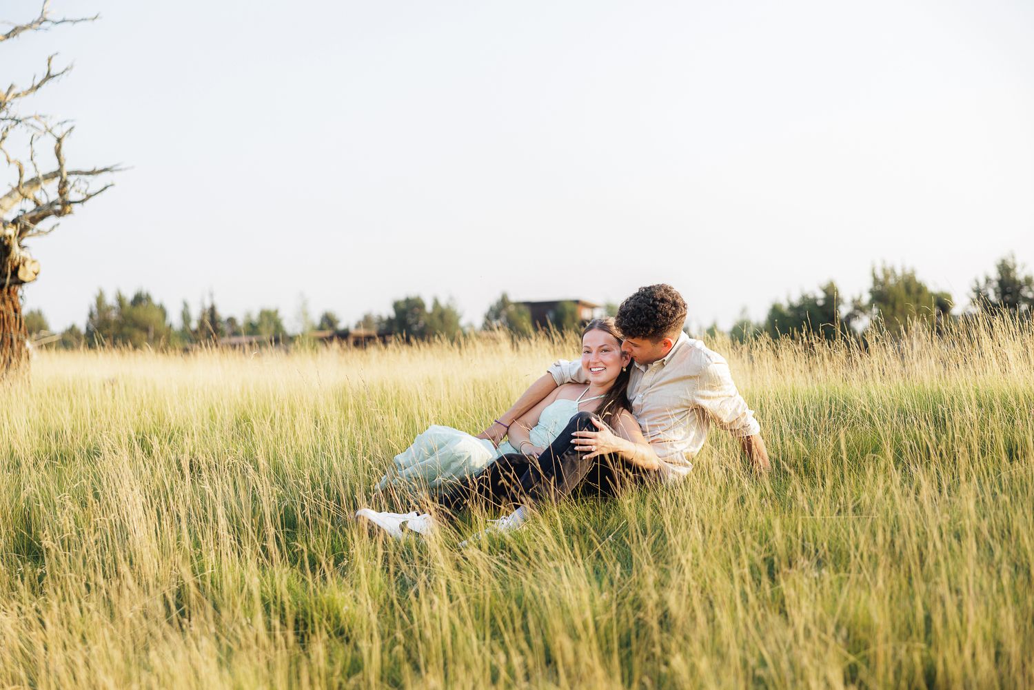Couple sitting together in tall green grass field, sharing a romantic moment outdoors on a sunny day.