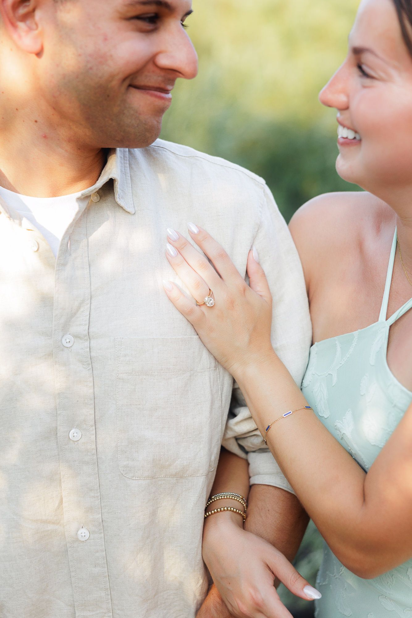 Couple smiling at each other, woman wearing engagement ring and bracelet, man in beige shirt, outdoor setting.