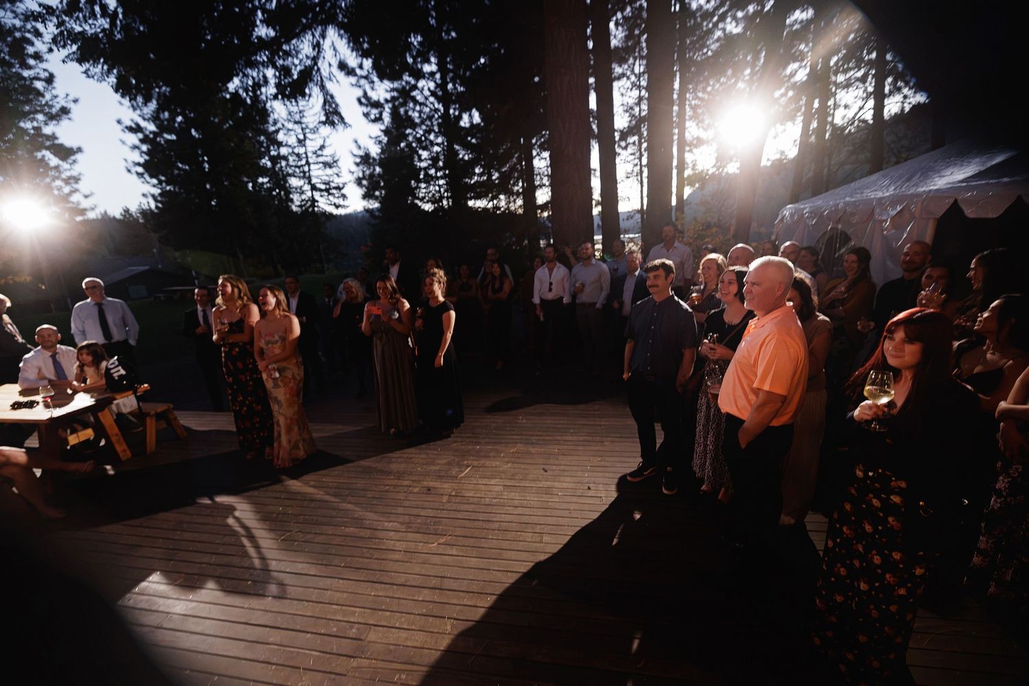 Guests gather on a wooden deck at dusk surrounded by tall trees at an outdoor evening wedding reception.