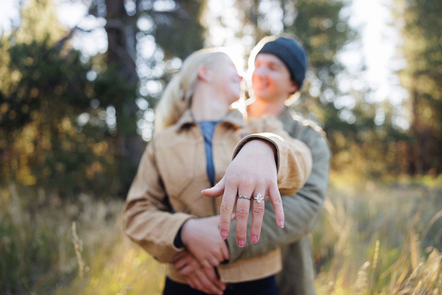 Couple embracing outdoors in a sunlit forest as the woman shows off her diamond engagement ring in the foreground.